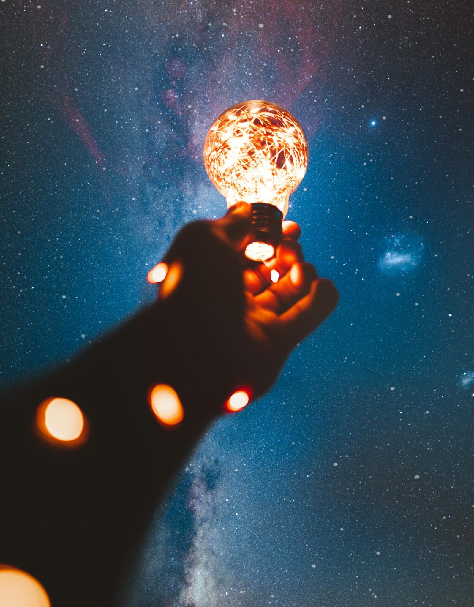 A hand holds a glowing bulb into the Milky Way night sky, creating a magical cosmic connection.