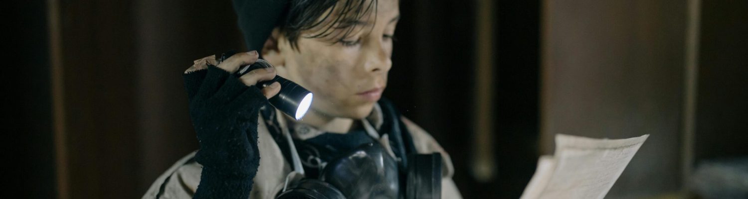 A young person in survival gear reads a paper using a flashlight in a dimly lit room.